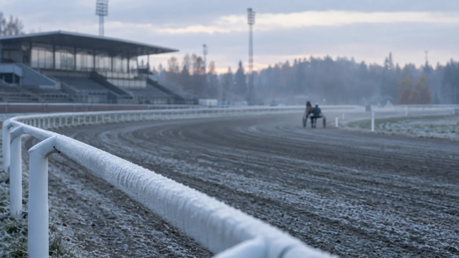 Frostig travbana tidigt på morgonen med ett räcke i förgrunden och en häst som joggar långt bort i oskärpa.