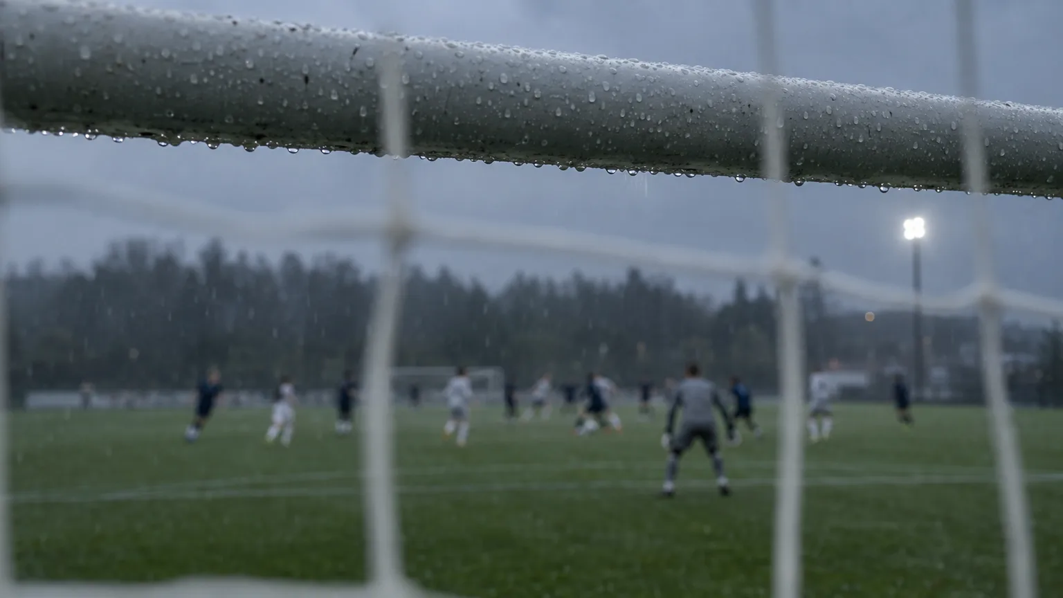 Fotbollsmål sett bakifrån nätet i duggregn, med oskarpa spelare långt bort i straffområdet under mulet kvällsljus.